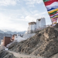 Scenic landscape of Ladakh: Majestic snow-capped mountains rise against a clear blue sky, with a winding river cutting through the rugged terrain. Traditional Tibetan-style architecture dots the landscape, offering a glimpse into Ladakh's unique culture and history.