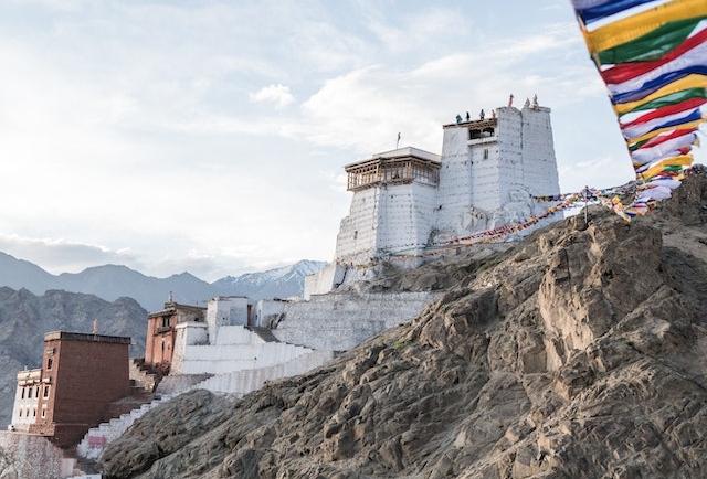 Scenic landscape of Ladakh: Majestic snow-capped mountains rise against a clear blue sky, with a winding river cutting through the rugged terrain. Traditional Tibetan-style architecture dots the landscape, offering a glimpse into Ladakh's unique culture and history.