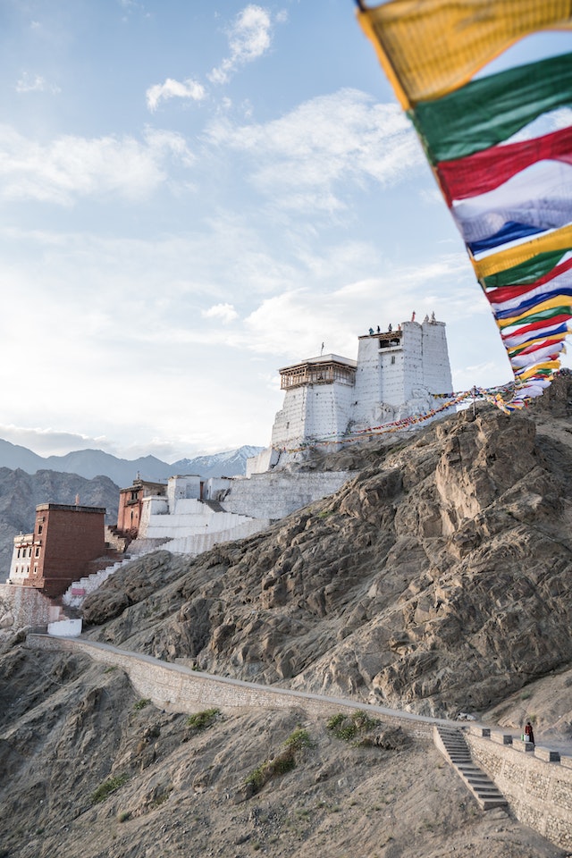 Scenic landscape of Ladakh: Majestic snow-capped mountains rise against a clear blue sky, with a winding river cutting through the rugged terrain. Traditional Tibetan-style architecture dots the landscape, offering a glimpse into Ladakh's unique culture and history.